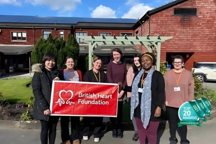 group-of-care-workers-and-staff-stood-outside-right-at-home-cheshire-east-with-a-british-heart-foundation-banner-celebrating