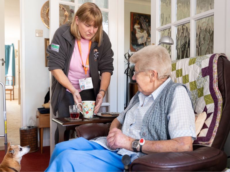 A carer gently handing a cup of tea to a seated senior at home, showing personalised support and companionship during daily living.